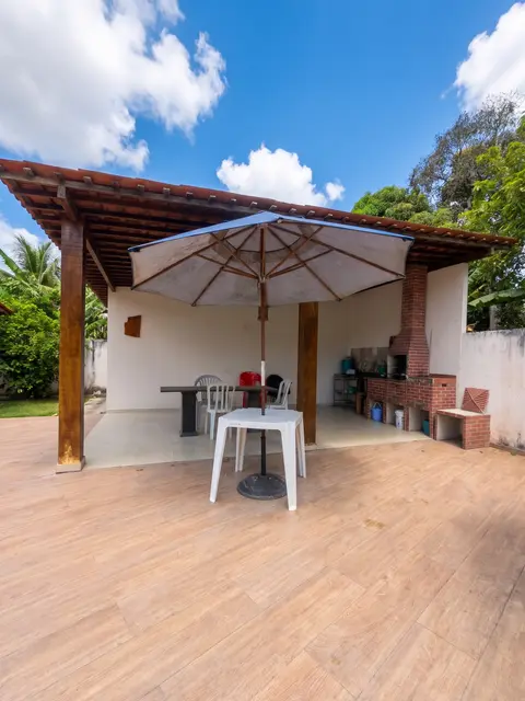 Área coberta com mesa e guarda-sol, churrasqueira ao lado e árvores ao fundo